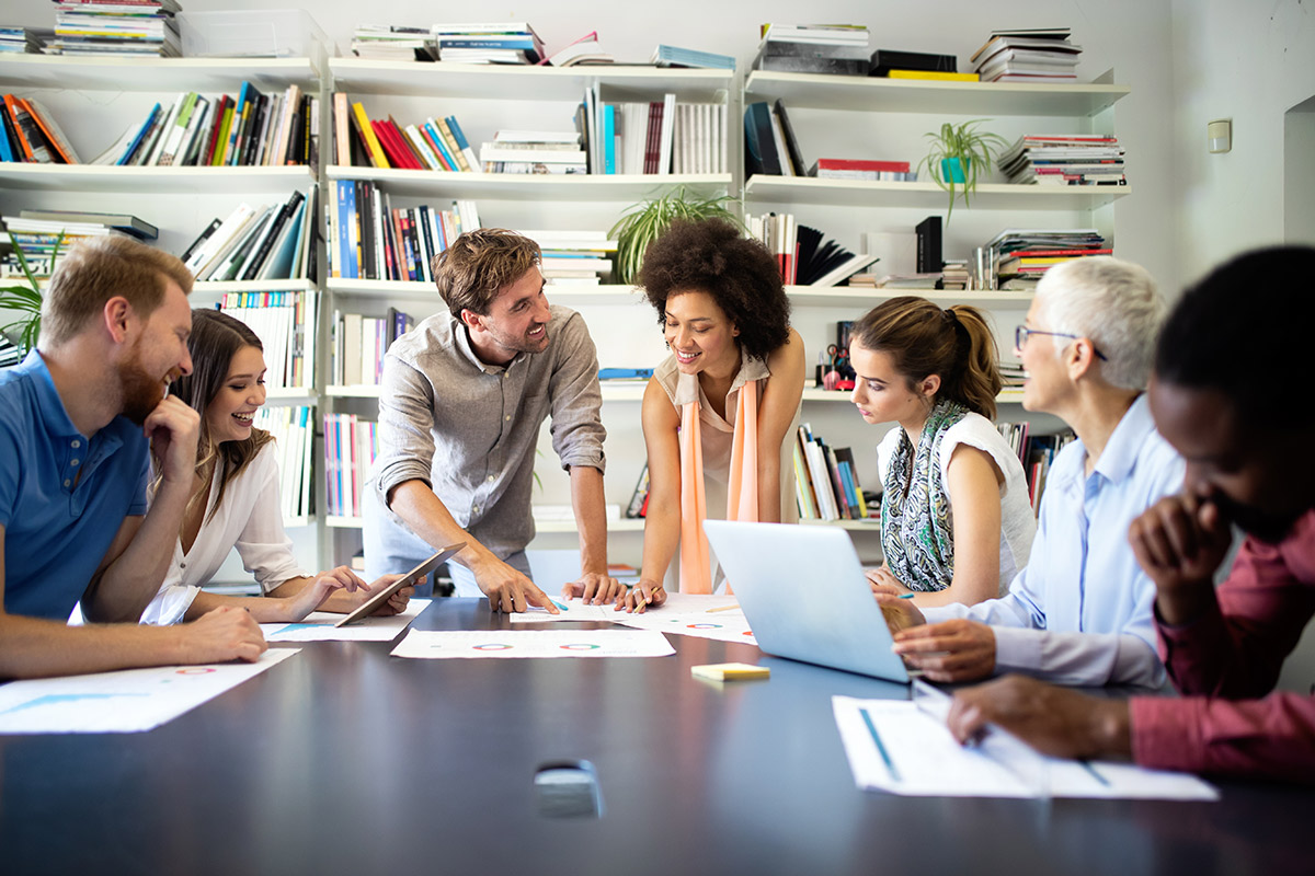 smiling employees gathered around a table to discuss educational assistance programs. | Corporate Synergies