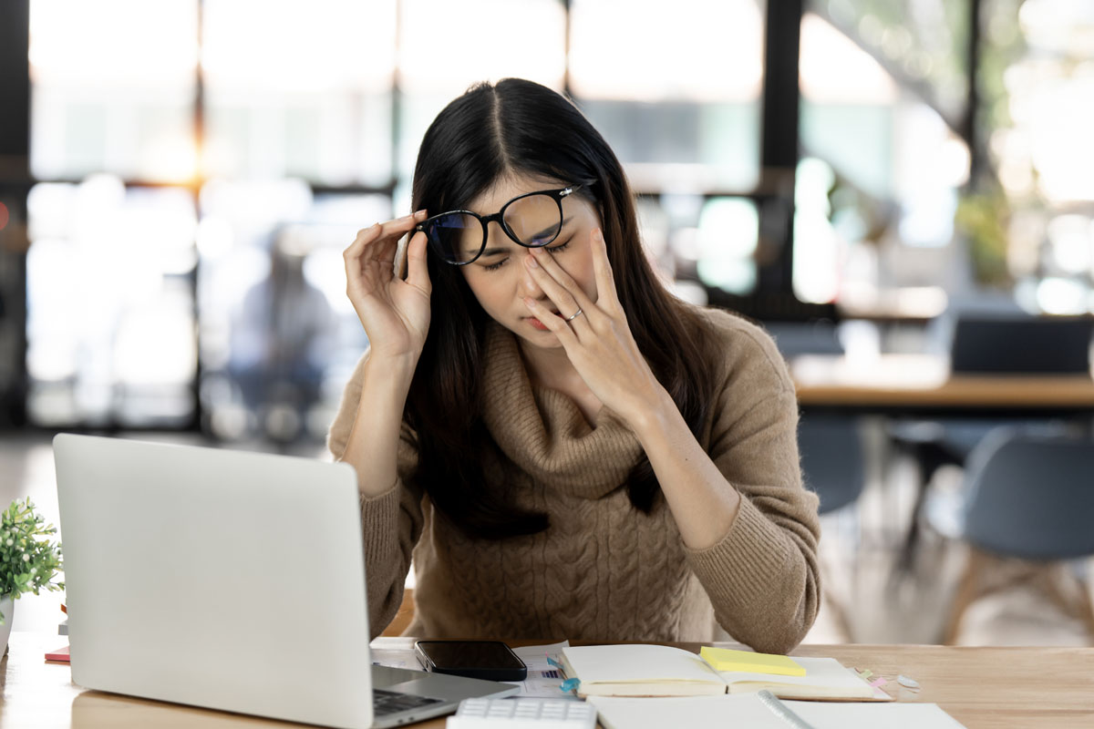 an example of workplace burnout: woman rubs her eyes at her cluttered desk | Corporate Synergies