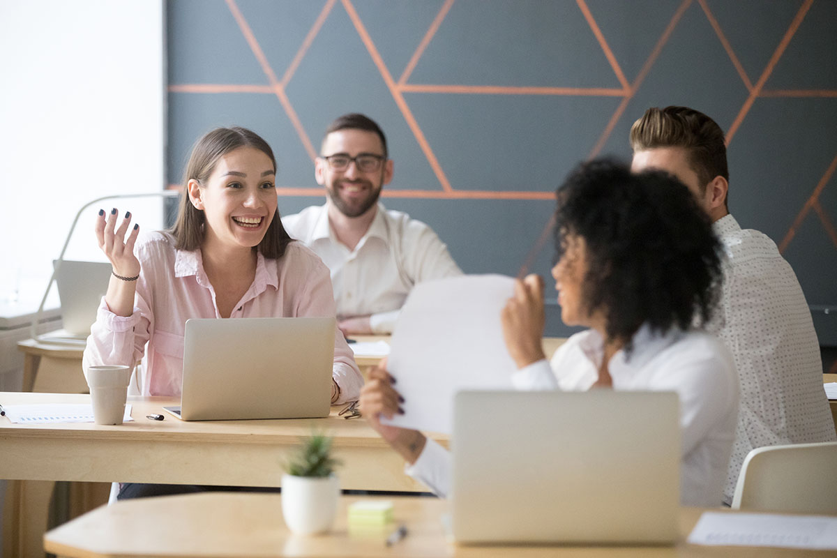 Employees sitting at tables with laptops discussing the benefits culture at work. | Corporate Synergies