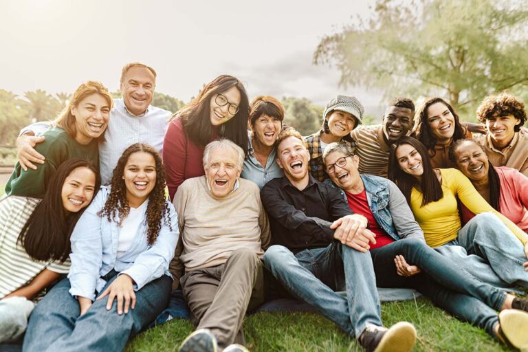 Blended sandwich generation family sitting in a park and laughing at the camera |Corporate Synergies